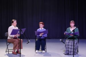 Three women sit in chairs and read from books dressed in pastel cardigans against a black backdrop.
