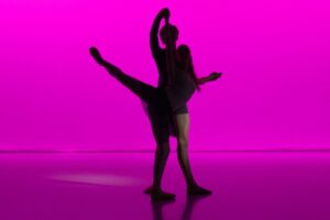 The silhouette of two dancers is shown in front of a bright fuschia backdrop. One female dancer is in an attitude back with her right leg with her left standing leg in a soft plie. Her right arm is held above her by a male dancer behind her, and her left arm is also held slightly behind her. The boy stands strong as she kicks her leg backwards.