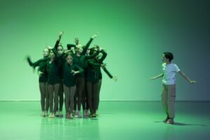 Against a bright green backdrop, a group of dancers on stage right form a clump with their arms bent in various angles and hands forming claw shapes. The dancers wear brown leggings and long-sleeved tunic tops with strands of small leaves braided into their hair. A boy wearing a white shirt and khaki trousers stands on stage right watching the group move. His arms are outstretched to the side, mid-exploration. He looks intrigued by the clump of figures.