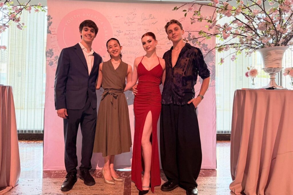 Millán, nanoha, samantha and ivan pose in front of a pink backdrop. Two tables with pink cloths hold a large vase of pink cherry blossom branches each, framing the four. Millán wears a dark black jacket and slacks with a white undershirt with the top button undone. Nanoha wears an olive green flowy dress with a bow around the middle. Samantha wears a form-fitting red dress with a high slit. Ivan wears a sheer black shirt with the sleeves rolled up and black wide-leg trousers.