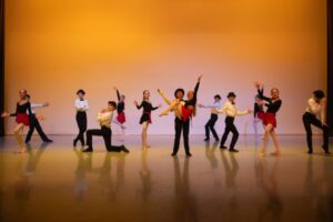 Boys dressed in white shirts, black trousers and top hats pose with girls dressed in black long-sleeved leotards, red skirts and pink tights. The backdrop is a warm orange, and the dancers are beaming in different poses involving jazz hands, kneeling, beveled feet, crossed arms, hat tips and pointed fingers.
