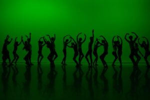 Silhouettes of dancers in front of a dark, vivid green backdrop are in a line across the stage. Each dancer stands in a different pose, involving parallel plies, arms raised in 5th, contracted torsos and beveled feet.