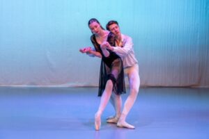 Eva and alex hold hands and curve their bodies. Alex is behind eva wearing a white flowy shirt with a red flower in the lapel, white tights and white shoes. Eva wears a long sleeve black lace leotard with a sheer knee-length skirt, pink tights and pointe shoes. Her hair is down, tied half up.