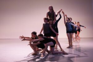 Dancers dressed in black shorts and grey tops create a dynamic tableau. At the front, two boys kneel on one knee and reach their arms forward in front of their faces. Behind them, a girl is lifted into the air, her left leg raised in an arabesque. Behind them, other dancers reach their arms up in a diagonal towards the girl.