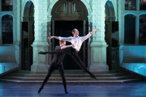 Harry, dressed in all black, lifts clovis in a sissone arabesque. Clovis wears a white button down shirt and black tights.