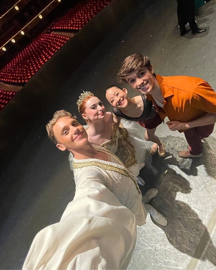 The four students take a selfie on stage in costume with the empty audience seats in the background. From left to right stand ivan, samantha, nanoha and millan.