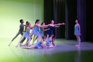Dancers dressed in shades of blue leotards with skirts and grey leotards and tights with white socks for the boys reach towards a single girl standing and facing the group. The backdrop is a light green colour that bleeds onto the stage floor. The girl stands tall in front of the group leaning and reaching towards her.