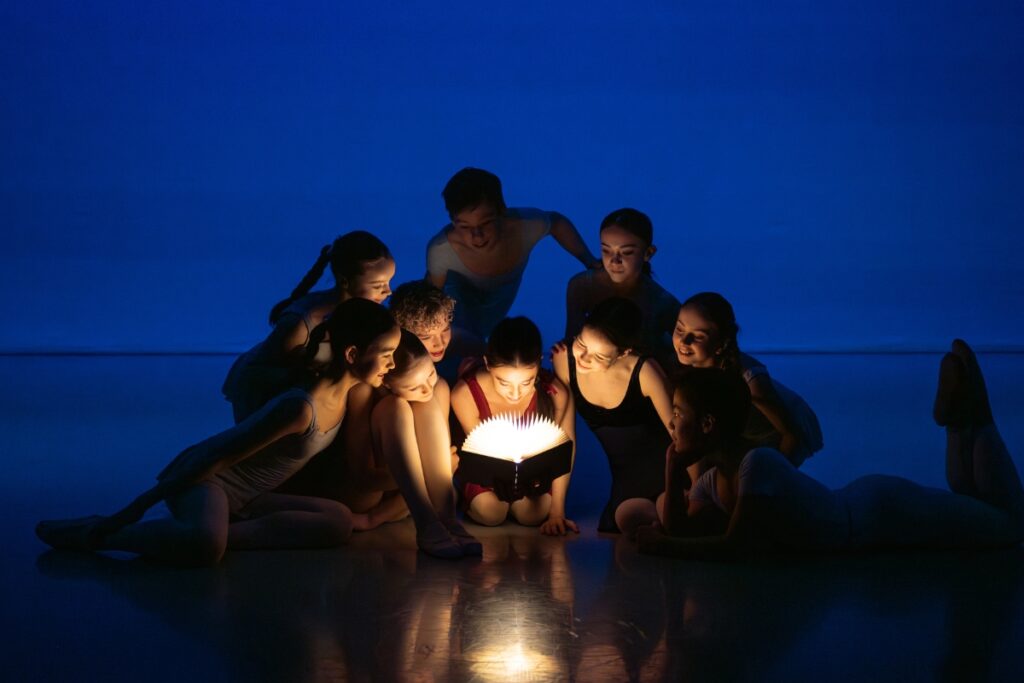 A group of dancers huddle together on the stage floor in dark lighting. The backdrop is a dark shadowy blue. The dancers' faces are illuminated by a glowing book, held by a young girl in the center of the group.