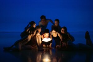 A group of dancers huddle together on the stage floor in dark lighting. The backdrop is a dark shadowy blue. The dancers' faces are illuminated by a glowing book, held by a young girl in the center of the group.