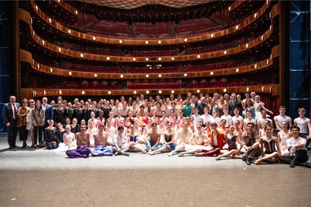 All the student performers pose in a group photo on stage with the empty audience in the background. They all wear costumes.