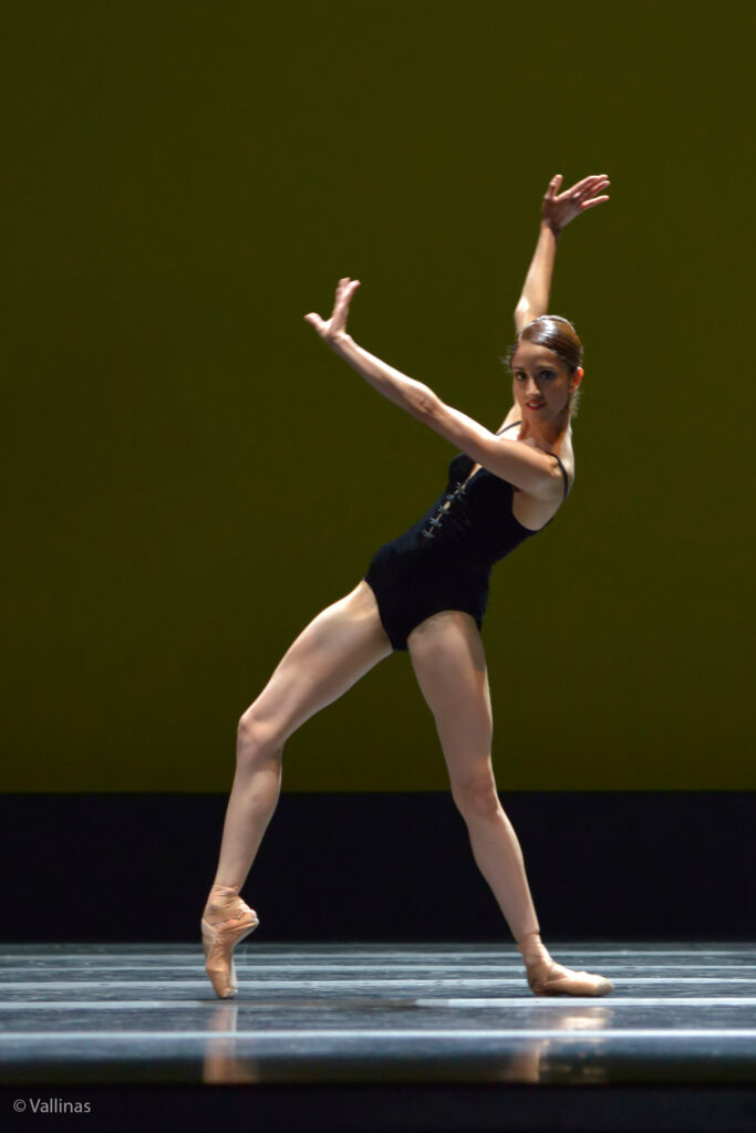 Nandita wears a black leotard with spaghetti straps and a corset-like closure on the front. She is in a contemporary pose, with her right leg bent and in forced arch to the front, her right hip jutted forward and her left leg leaning forward on a diagonal with her foot flat on the floor. Her hands are flexed, with her left arm crossing in front of her and her right hand above her head to the back. Her hair is slicked into a french twist.