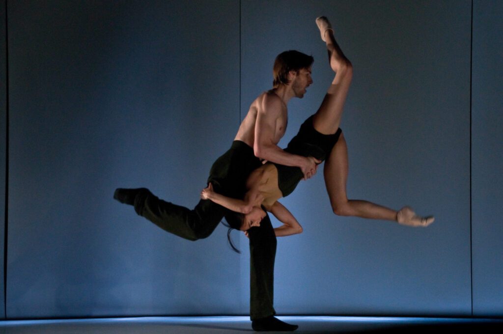A male dancer, shirtless with black trousers and socks, balances on a bent left leg with his right leg back in a parallel attitude position. Nandita holds onto his thighs upside down. He holds onto her waist as her legs are in attitude positions in the air in front of him. They both face stage left. Nandita wears a sleeveless black leotard and shorts. They are in front of a dimly lit blue backdrop.