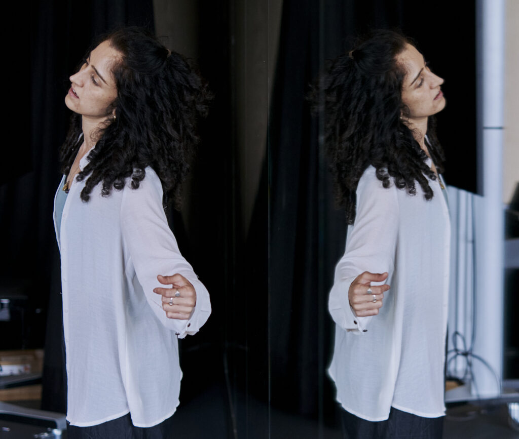 Nandita stands with her back to the mirror, her reflection on the other half of the image. She wears an unbuttoned white button down shirt, loose and flowy, and stands with her arms gently outstretched to the sides with her back slightly arched. She looks towards the dancers rehearsing out of frame. Her long curly hair is half up half down.