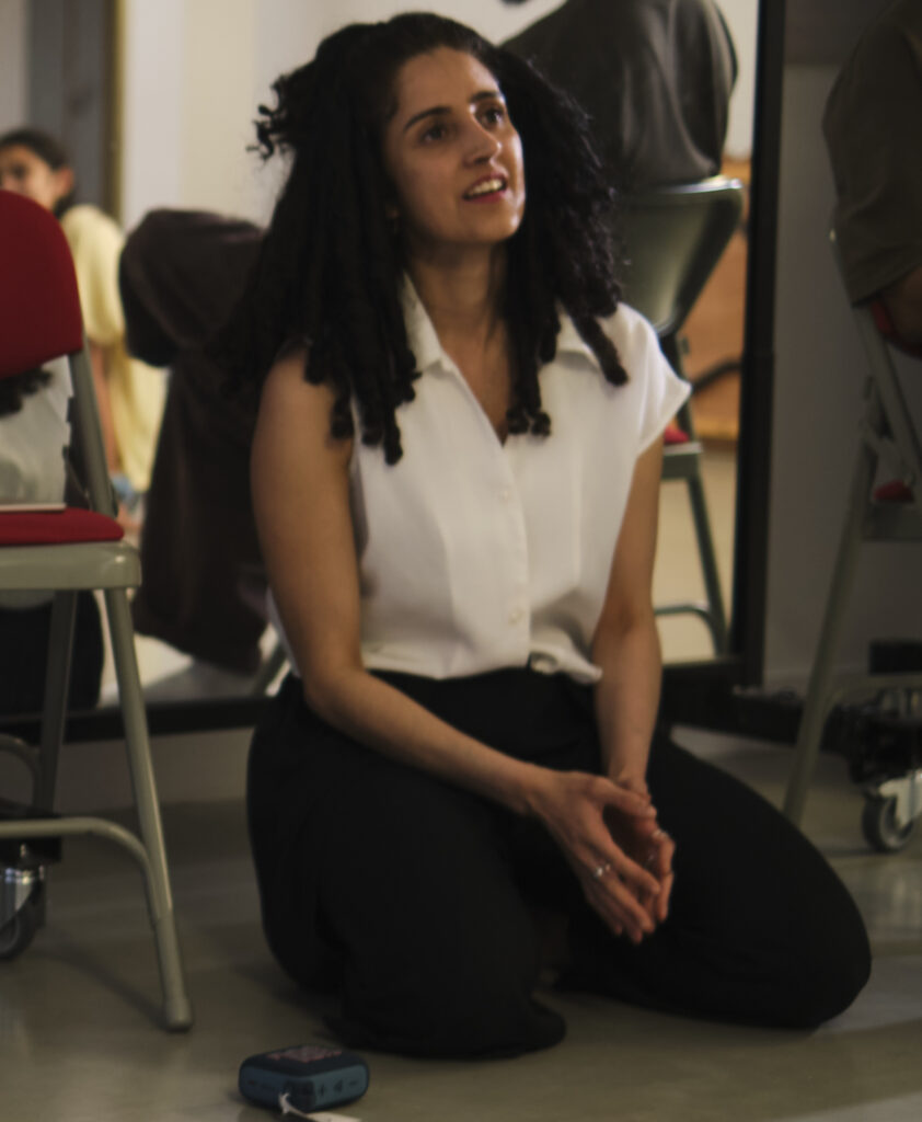 Nandita sits in a chair in front of a mirror watching the dancers. She wears a white sleeveless shirt and black trousers. Her hair is down to her shoulders, and her mouth is in a soft smile, slightly open as if to say something.