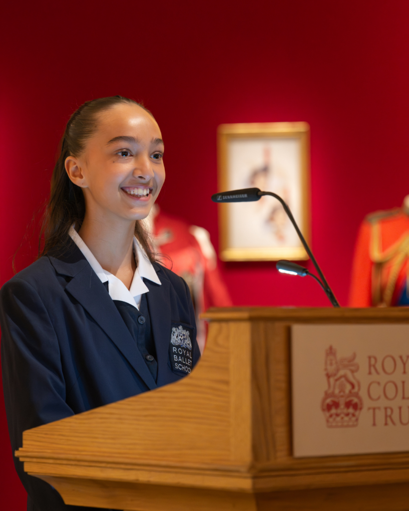 Nelia stands behind the wooden podium, wearing her school uniform and smiling to the crowd.