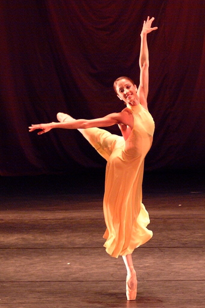 Nandita wears a long yellow dress on stage. She is in an arabesque with her left leg back and standing on her right leg on pointe. Her left arm is raise in an elonge above her head, and her right arm is extended to the side.
