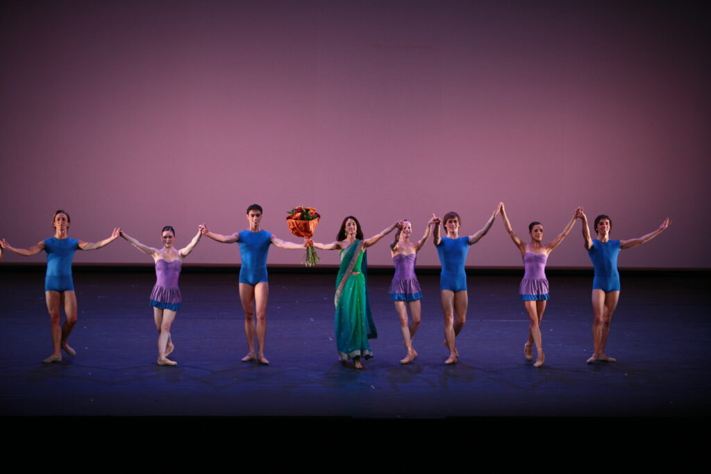 Nandita stands center stage wearing a green dress alongside seven dancers who performed in her piece capture. The three female dancers wear purple and blue short ruffled dresses with fitted bodices. The four male dancers wear cobalt blue unitards with shorts. Nandita holds a large bouquet of orange flowers. They hold hands just before bowing.
