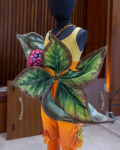 A black mannequin wears a unitard with tank sleeves. It is yellow on the top from the mid chest up and orange on the bottom and trousers. A large prop of leaves and an oversized blackberry cover the front of the costume.