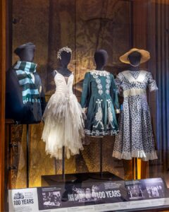 Four black mannequins display uniforms and costumes from the past. From left to right: a navy blue blazer with the royal ballet school crest and a scarf wrapped around the neck with light blue and dark green stripes; a little swan dress with a white crown and feather details; a dark green traditional irish dance dress with intricate white patterns running down the front symmetrically; a white dress with a blue floral pattern, a white lace collar and a straw hat.