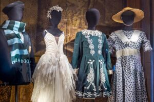 Four black mannequins display uniforms and costumes from the past. From left to right: a navy blue blazer with the royal ballet school crest and a scarf wrapped around the neck with light blue and dark green stripes; a little swan dress with a white crown and feather details; a dark green traditional irish dance dress with intricate white patterns running down the front symmetrically; a white dress with a blue floral pattern, a white lace collar and a straw hat.
