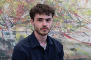 Jake stands in front of a backdrop with colourful muted brush strokes. He wears a black polo shirt with a white undershirt. He has short brown hair and some facial hair.