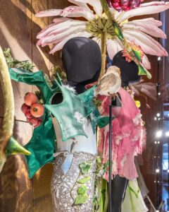 A close up of the winter fairy costume: a light grey unitard is covered by darker grey sparkly mesh on the bottom half. On top of the chest and over the shoulder along the back are oversized holly leaves with large red berries on the back. The pink dress and large flower prop can be seen in the background.