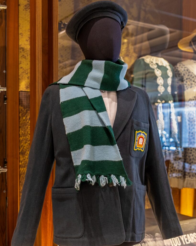 The evolution of the royal ballet school crest A navy blue blazer with the royal ballet school crest and a scarf wrapped around the neck with light blue and dark green stripes on a black mannequin.