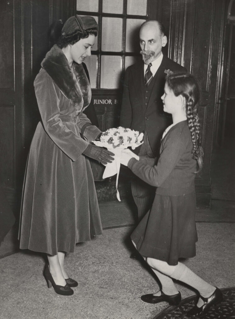 100 stories - arnold haskell Princess margaret wears a long fur-trimmed coat and heels and takes a bouquet of flowers offered to her by a young girl in braids and a dark dress. Arnold stands in the background watching the exchange.