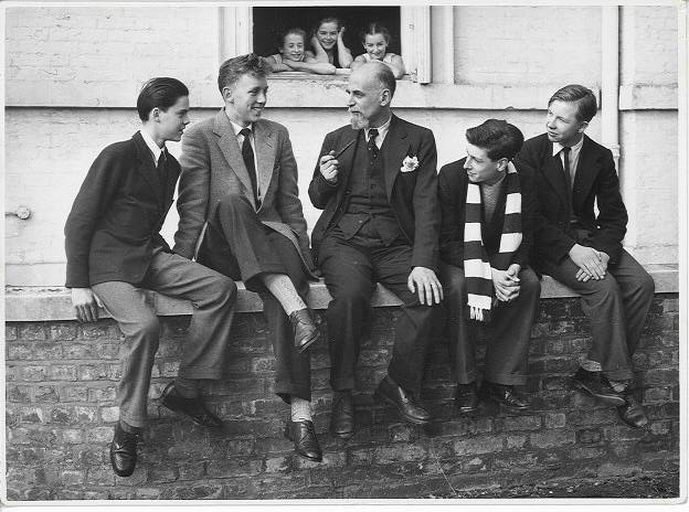 100 stories - arnold haskell Arnold haskell sits on a low brick wall with a group of young students at white lodge in 1955. All of them wear suits and ties. Arnold sits in the middle with two boys on either side. In a window in the background just behind arnold's head, three female students poke their heads out to listen.