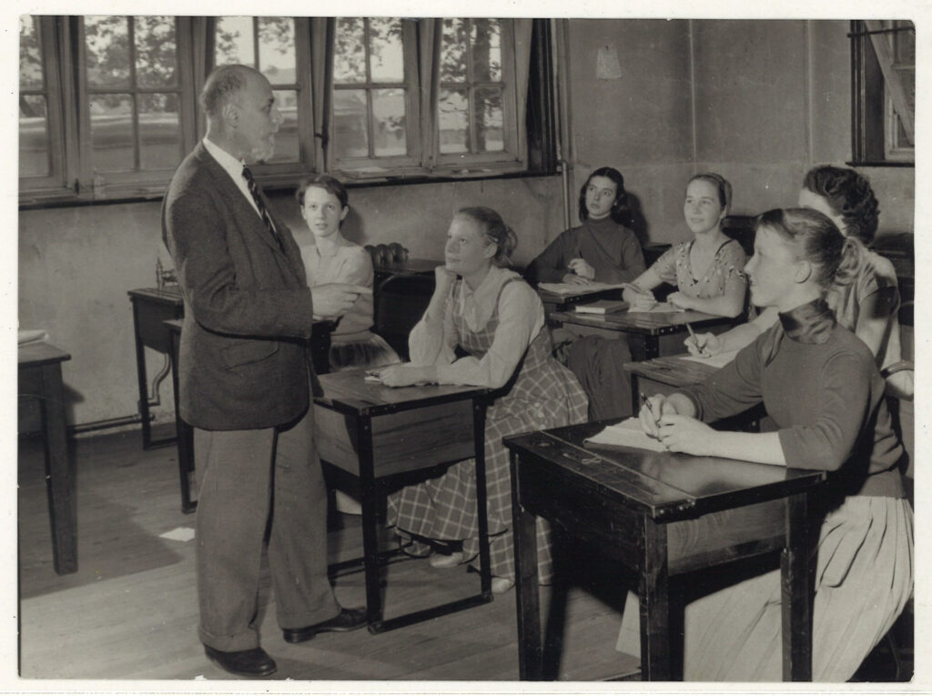 100 stories - arnold haskell Arnold faces a class of young female students sitting in desks and wearing casual dresses.