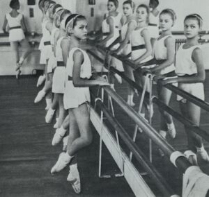 Leslie stands at the barre in the foreground facing the mirror with her head turned towards the camera on her right. She wears a short white tunic belted at the waist, a white headband, white socks and ballet shoes. The photo is greyscale.