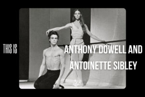 Anthony kneels on both legs sitting back on his feet wearing black tights with no shirt. He faces downstage right with his head towards the audience. His arms hang by his sides. Antoinette stands beside him, one hand on a barre and the other a la seconde. She wears a loose white tunic with her hair down. She also faces the audience.