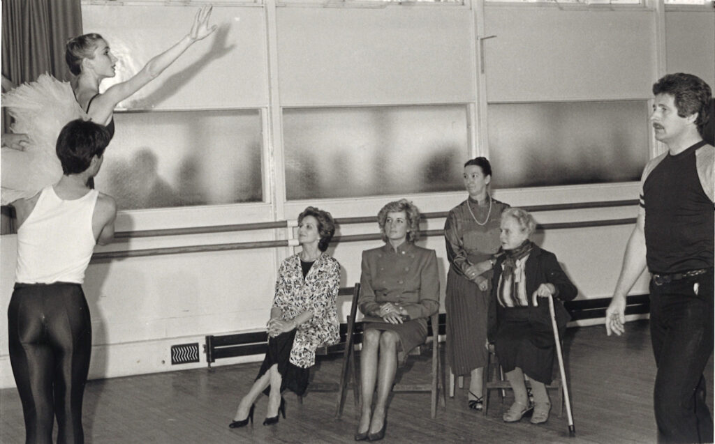100 stories: barbara fewster On the far left of the image, a young boy lifts a young girl in a tutu on his shoulder. Seated in front of them and looking up at the girl are merle park, princess diana, barbara fewster and ninette de valois. David drew stands to the far right, walking towards the students.