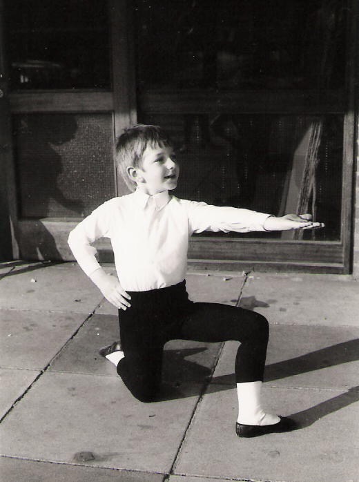 100 stories: christopher saunders Christopher, aged five, kneels on his right knee with his right arm on his hip and left arm extended forward. He looks to his hand and smiles. It is a black and white photo.