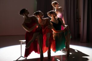 Four female dancers are illuminated from the back by a white spotlight on a dark stage. Three sit on a bench with their left legs crossed over their right, both feet pointed in pointe shoes. They wear a red, orange and green flowy dress with a v-neck and short sleeves. Their hair is swept into french twists, and they lean to the right while looking up to the left. Their hands are crossed on their knees. A fourth dancer in a pink ombre dress stands just behind the girl on the left of the bench, also leaning to the right and looking up to the left.