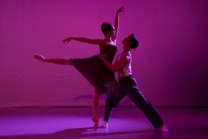 The scene is tinted in a deep purple-pink light. A female dancer balances in arabesque in a black sleeveless dress, her left arm extended above her and her right hand to the side. A male dancer lunges towards her holding her waist. He wears a white sleeveless shirt and black trousers.
