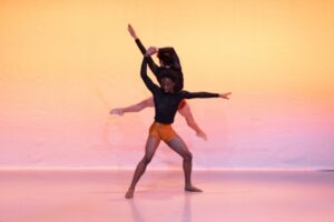 Two male dancers wear long-sleeved black mesh shirts and orange shorts. One dancer stands in a wide parallel second position, left arm extended to the side and right arm bent above his head, holding the right hand of the other dancer behind him. The other male dancer is mid-jump facing backstage, his back touching the first dancer's shoulders and upper back. His legs are in a wide second position, feet pointed, and his left arm is extended up to the left.