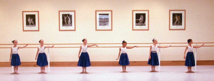 100 stories: barbara fewster A line of young girls in white leotards and long solid blue skirts stand in front of the barre in the upper school studio. Their feet are in first position in skin-tone shoes and bare legs. They look to the left as they raise their left arms limply to the side. Above the barre, photographs of notable alumni are framed in wood that matches the barre and baseboards.