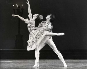 Leslie poses in arabesque with her arms extended forward. She looks straight up, with a large tiara perched on her head. Her white tutu has dark stripes and zig zags. A male dancer holds her waist from behind, in a lunge with his right leg forward. He follows her eyeline, looking upwards. He wears a matching white top with long sleeves and dark designs with white tights and shoes.