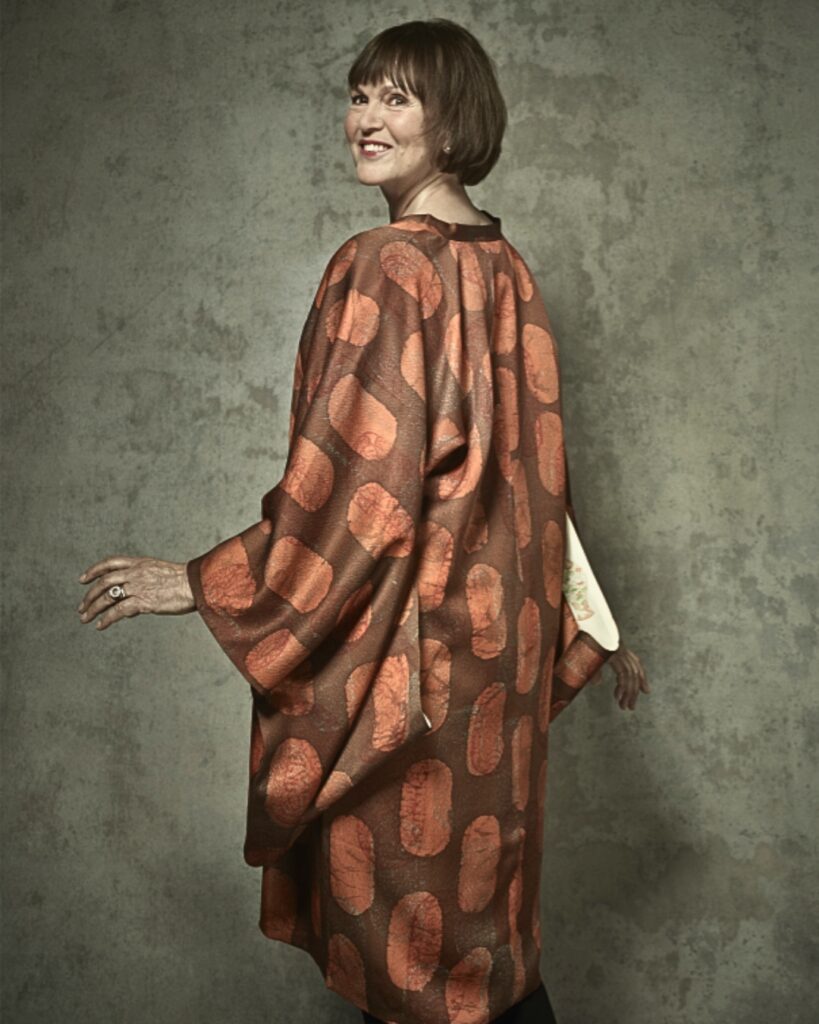 Leslie wears a long loose brown orange dress against a grey dappled backdrop. Her hair is in a short bob with fringe. Her back is angled to the camera and she turns her head over her left shoulder to smile at the camera.