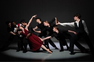 A female dancer in a deep red dress leans far to the side on pointe, held above the floor by three male dancers with four more male dancers trailing behind her, arms reaching towards her. The male dancers wear black trousers with a mix of white long-sleeve and black short-sleeve tops. The stage is dark, with the group illuminated by a white spotlight.