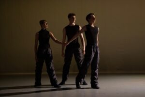 The stage is dimly lit, and a yellow-brown muted light illuminates the backdrop. Three dancers stand in a triangle facing downstage left. All wear black sleeveless tops with baggy black trousers and socks. A male dancer forms the point of the triangle, standing on his left leg with his right leg softly extended to the back, his toes still on the ground. He reaches his right arm back to hold the left hand of a female dancer. She stands on her right leg with her left leg extended forward, foot flat on the floor. Another male dancer stands behind both in a wide second position just behind their arms. His arms hang loosely to the side. All look towards the downstage corner.