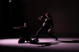 A dark stage is illuminated with a soft wide spotlight over two dancers. Both wear black suits and socks with a white undershirt, like business people. The female dancer is downstage right, kneeling on both knees with her right knee slightly more forward and her left leg slightly extended back. Both arms reach energetically forward, but her torso stays upright. The male dancer stands just behind her in a wide, off-kilter lunge with both legs bent, left leg forward. His torso leans back but his head tilts towards the woman. His arms are bent forward, his right arm held by his shoulder and left arm down by his hip.