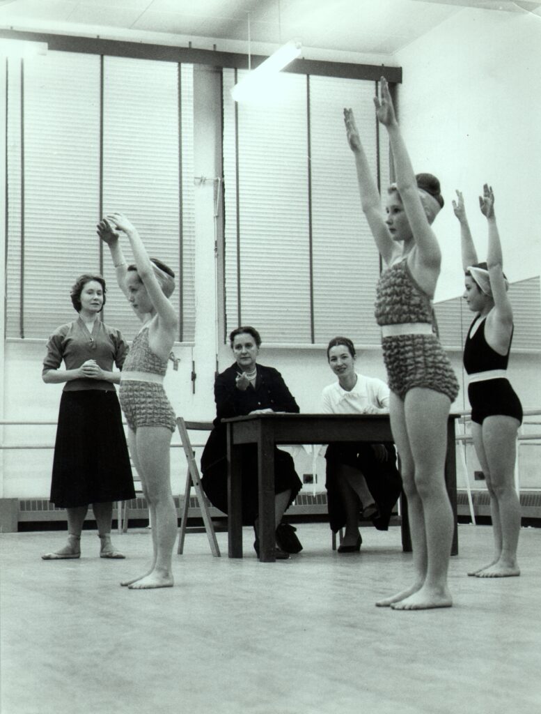 100 stories: barbara fewster Three young girls stand in the studio with their arms straight up. They wear textured leotards with large white strips around the waist. They are bare-legged and barefoot in parallel. Barbara fewster stands in the background with her hands together, watching the girls. She wears a short-sleeved blouse and long black skirt with ballet shoes. Ursula moreton and audrey harman sit next to her at a table, also watching the girls.