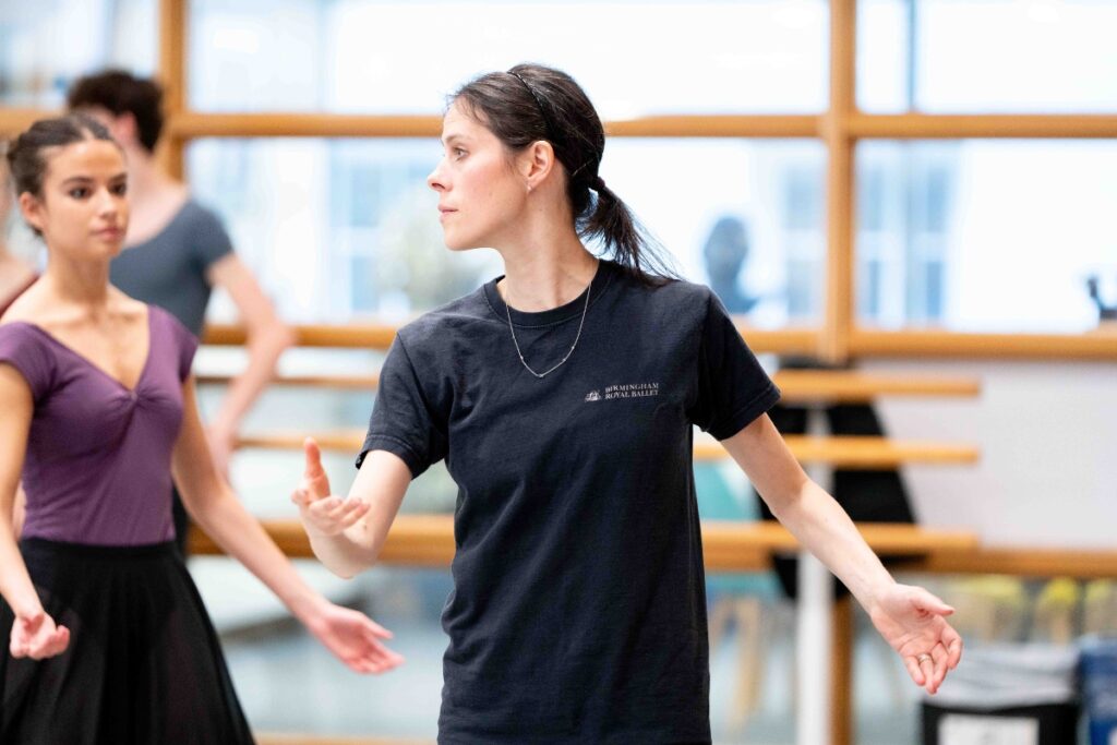 ‘refine all the time:’ alum samara downs on staging checkmate and swan lake for our summer performances Samara downs has her dark short hair tied back in a ponytail and wears a long black t-shirt with 'birmingham royal ballet' written on the front left. Her arms are gesturing in front of her, and her head is turned to the right in profile, looking towards the students.