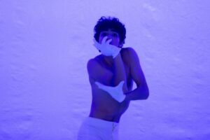 Blue light filters the stage and dancer. The male dancer wears white trousers and no shirt with short white gloves. He twists his torso toward the camera, left arm bent 90 degrees across his chest and right elbow bent up, with his hand curving around his chin and the front of his face.