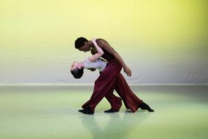 Green-yellow light illuminates the backdrop and the floor. Two dancers pose intertwined center stage. The male dancer faces stage right and curves his body over his left leg, extended to the front with his foot flexed and heel on the floor. His right leg is bent slightly to the back. He wears a black vest and red trousers. A female dancer holds onto his neck and leans backwards in front of him. Her back arches and her head gazes upwards. Her legs are extended front and down to the floor, with her feet on a diagonal releve. She wears a white vest and red flowy trousers.