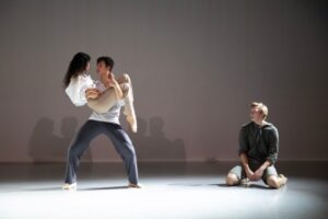 Two dancers are centre stage with one dancers offset to stage left. Centre, a male dancer stands in a parallel second position with a slight plie, holding a female dancer with his left arm under his knees and his right arm around her waist. She holds her left arm around his neck and touches his right hand with her right. They gaze intently at each other. Her wavy dark hair falls loosely down her back. She wears an oversized, long-sleeved white shirt and beige trousers and socks. He wears a white long-sleeve shirt, grey loose trousers and beige socks. The male dancer offset from them kneels widely on the floor, arms falling to the floor in front of him. He is slightly hunched and dejectedly watches the couple.