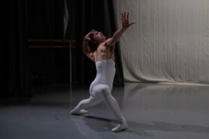 The stage is softly lit in a cool white light. A male dancer wears white full body tights that stop at the upper chest with skin tone suspenders over his shoulders. He is in a deep lunge with his right leg forward, bent and very turned out. His left leg is extended long to the back, foot flat on the floor. His left arm bends to grasp the back of his head, and his right arm is extended powerfully to the front, hand spread wide.