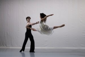 A male dancer lunges slightly to the back, extending his right arm downstage to hold the hand of a female dancer mid grand jete, her legs in an oversplit. The female dancer wears a flowy white dress with her dark brown hair flowing behind her in waves.
